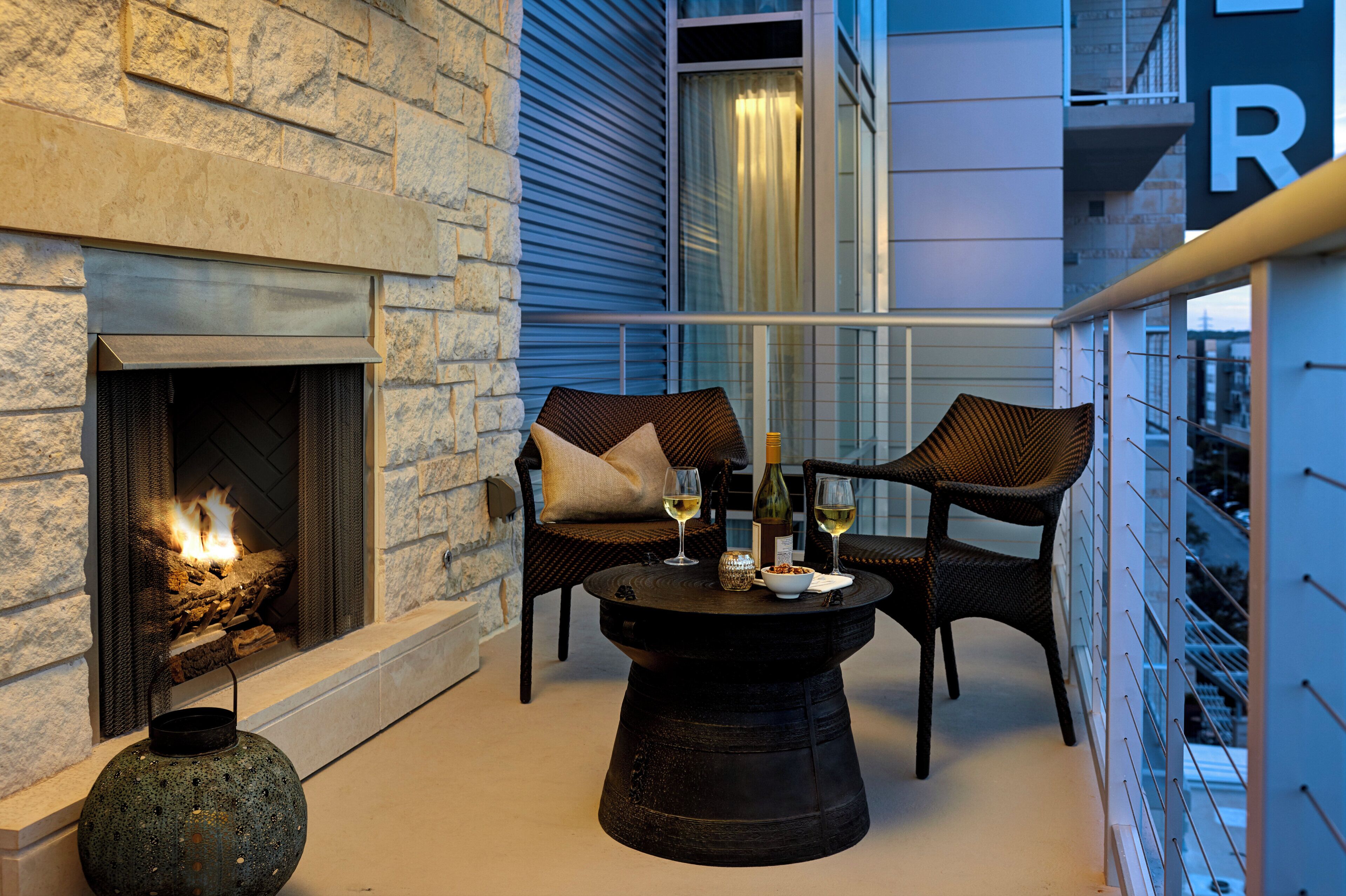 A balcony with a stone fireplace, wicker chairs, a round table with wine glasses and bottles, and a decorative lantern.
