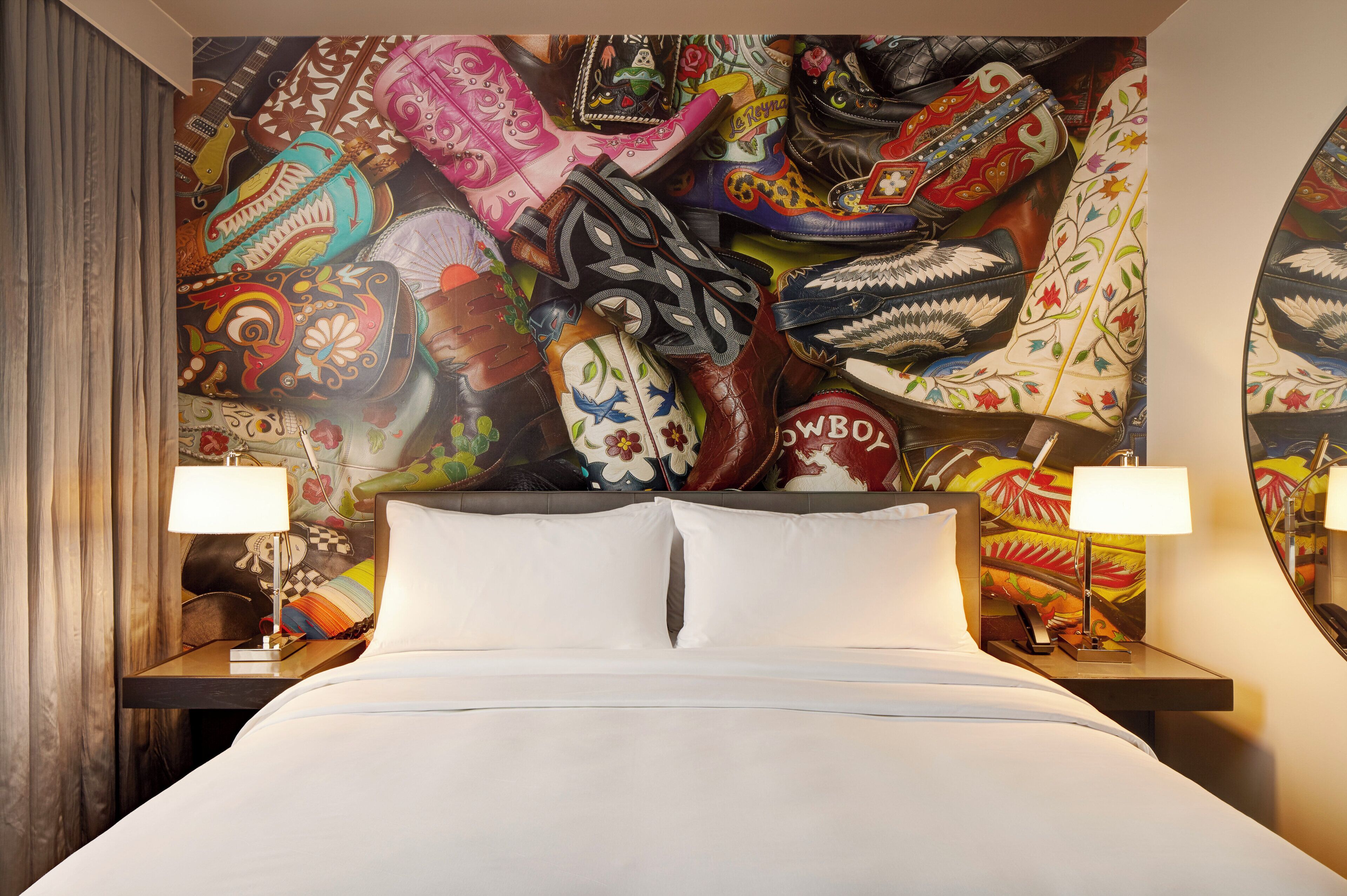 Quarto de hotel com uma cama grande, duas mesas de cabeceira com luminárias, e um mural vibrante com diversos padrões e designs.