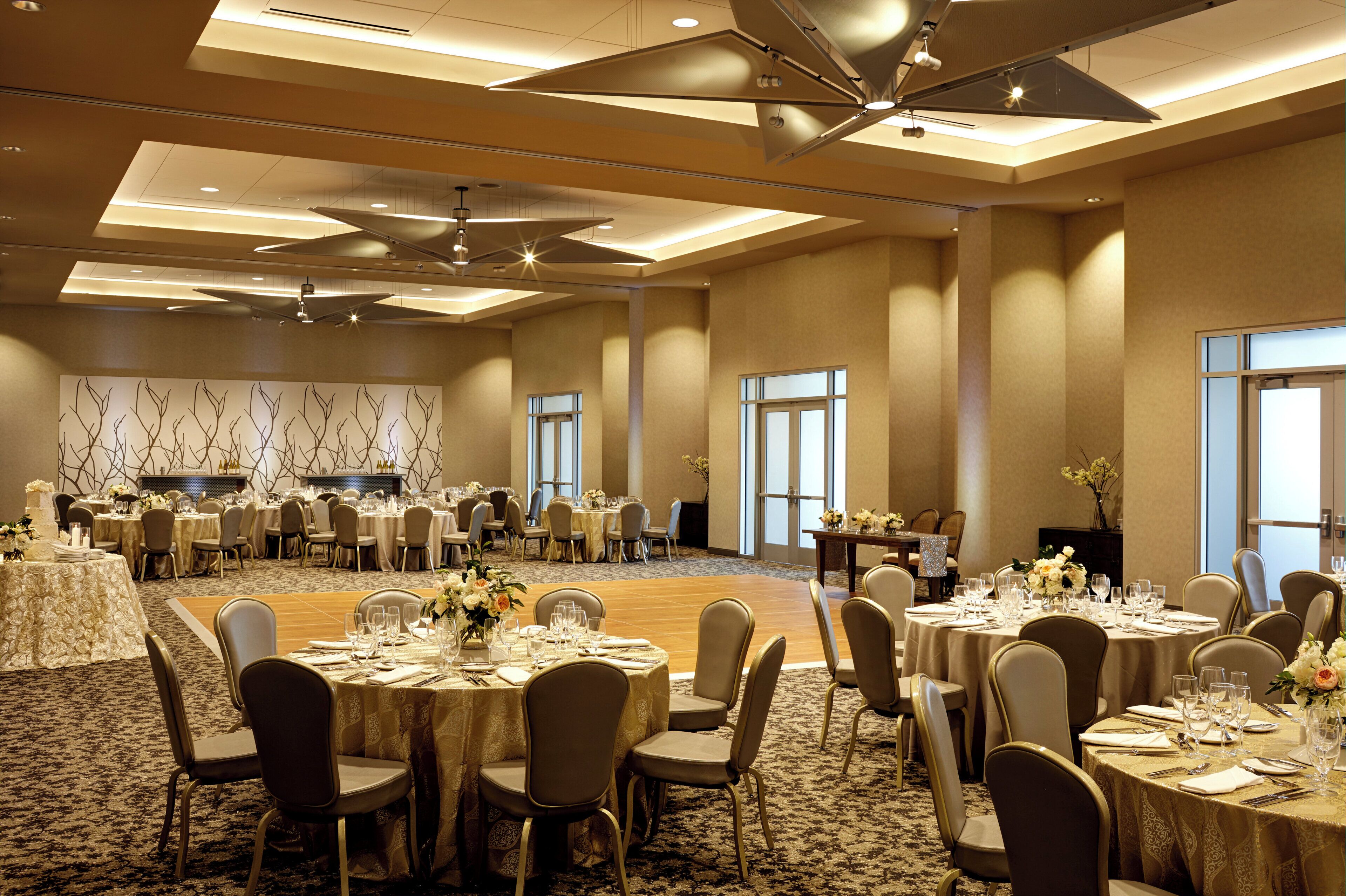 A large banquet hall with round tables set for dining, a stage with a backdrop, and a ceiling fan.