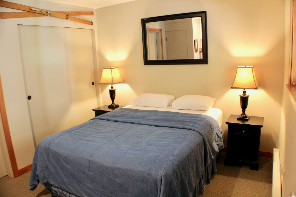 A neatly made bed with a blue bedspread, two bedside tables with lamps, a mirror above the bed, and a door on the left.