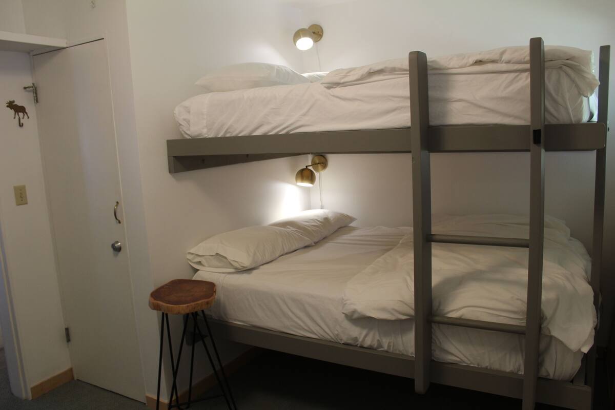 A bunk bed with two levels, a small stool, and a wall-mounted light fixture.