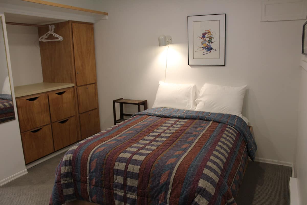 A bedroom with a bed, a wardrobe, a bedside table, and a wall-mounted lamp.