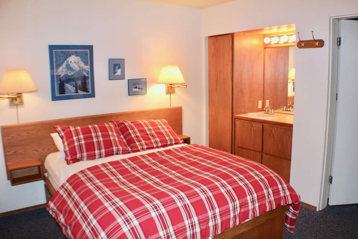 A bedroom with a bed, two bedside lamps, a wooden headboard, a wooden wardrobe, and a bathroom with a mirror and sink.