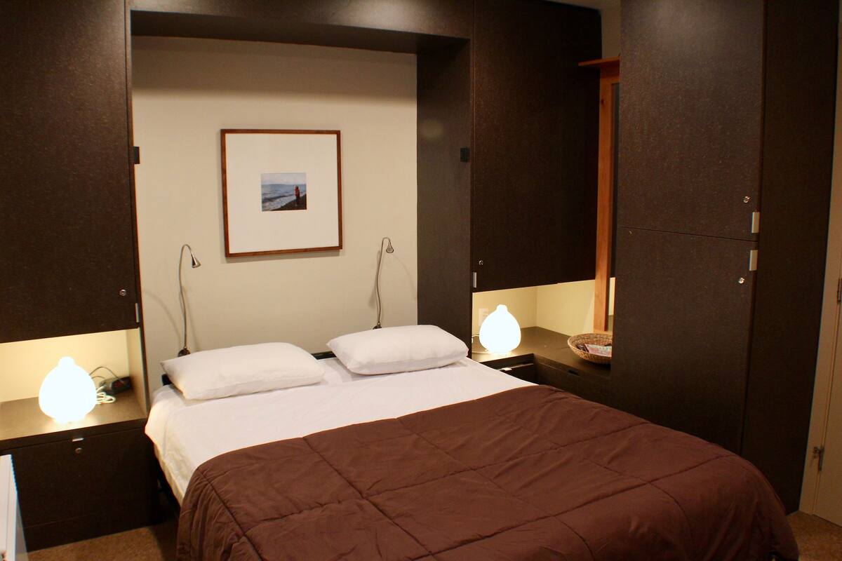 A neatly made bed with a brown blanket, two bedside lamps, a framed picture on the wall, and a wooden wardrobe.