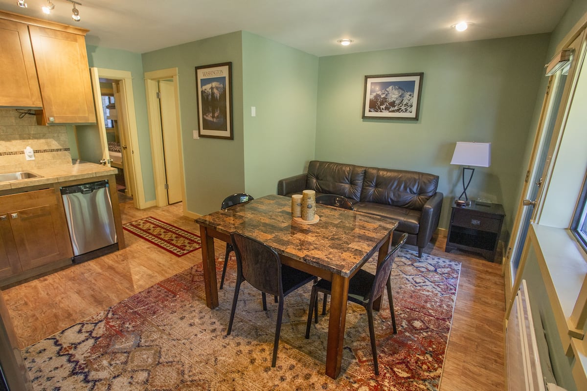 A compact kitchen with a dishwasher, a dining table with chairs, a leather sofa, and a rug.