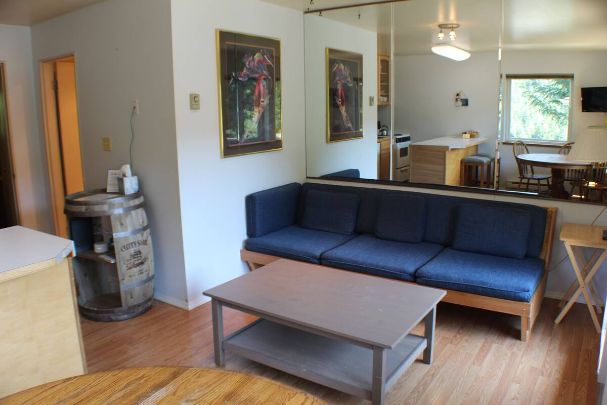 A living room with a blue sofa, a wooden coffee table, and a kitchen area visible through a glass door.