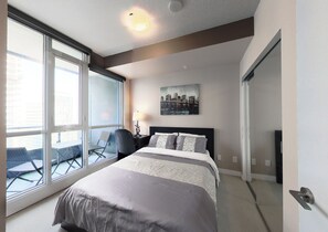A modern hotel room with a large bed, a desk with a chair, a cityscape painting, and a sliding door. - Spectacular Views Apartments (Toronto)