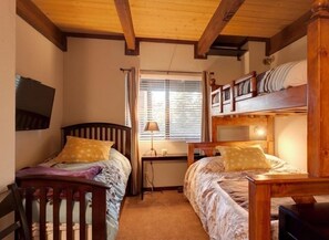 A bedroom with two wooden bunk beds, a desk, a chair, a TV, and a window with blinds.