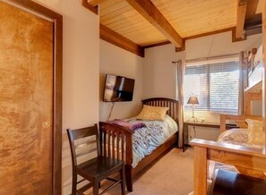A bedroom with a wooden bed, a TV mounted on the wall, a chair, and a window with blinds.