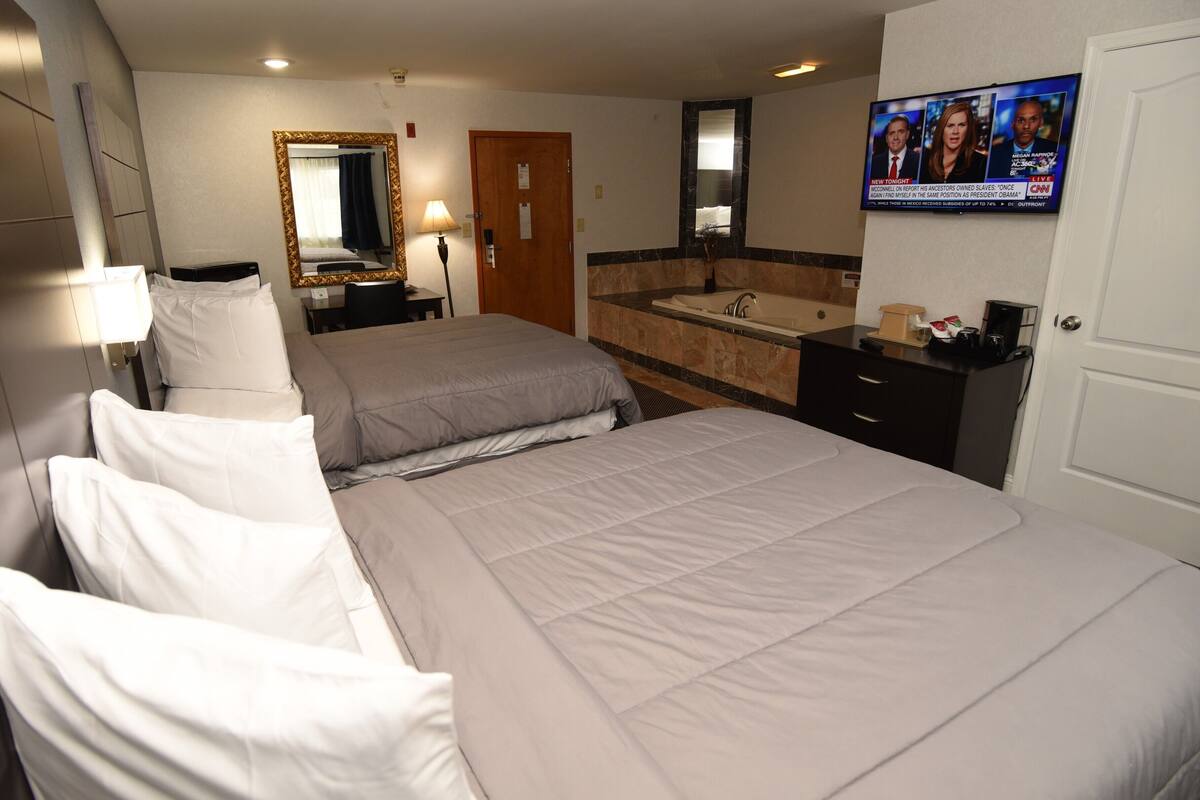 A hotel room with two beds, a bathroom with a sink and mirror, and a TV displaying news.