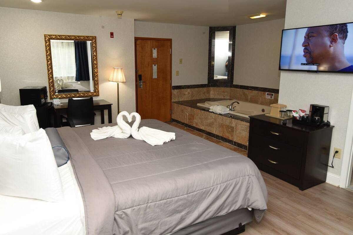 A hotel room with a bed, a desk, a mirror, a TV, a bathroom with a bathtub, and a coffee machine.