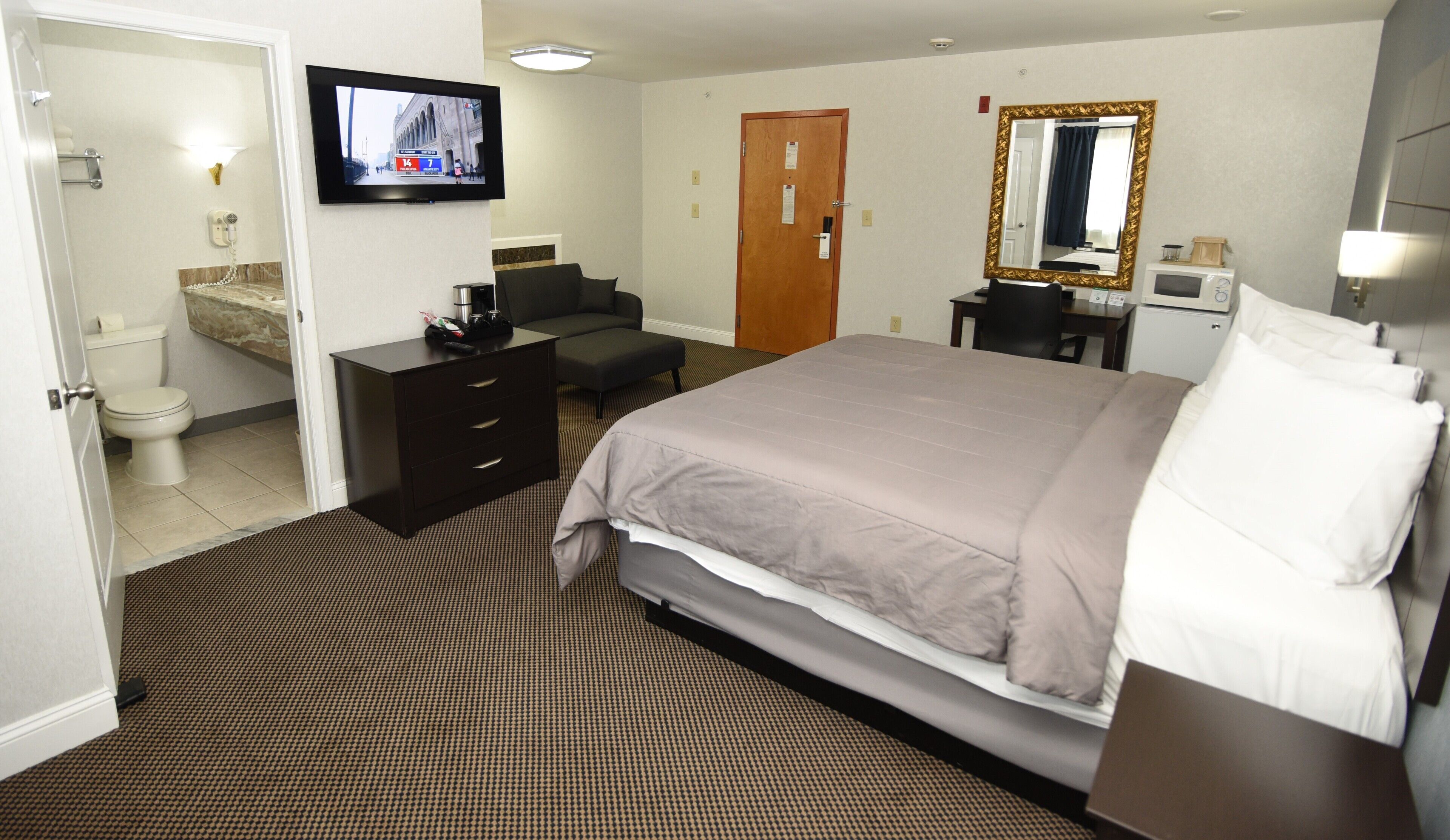 A hotel room with a bed, a desk, a chair, a television, a mirror, and a bathroom.