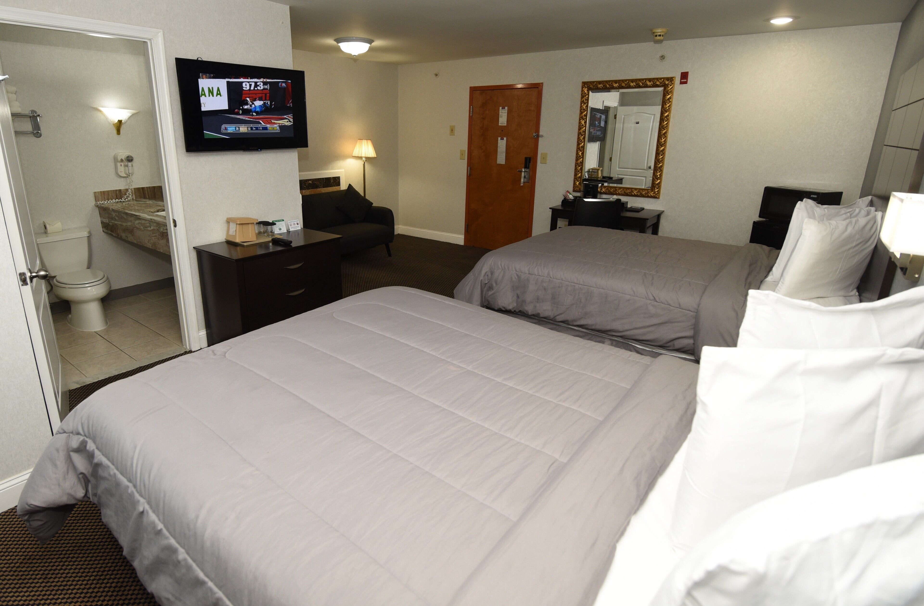 A hotel room with two beds, a TV, a desk, a mirror, and a bathroom.