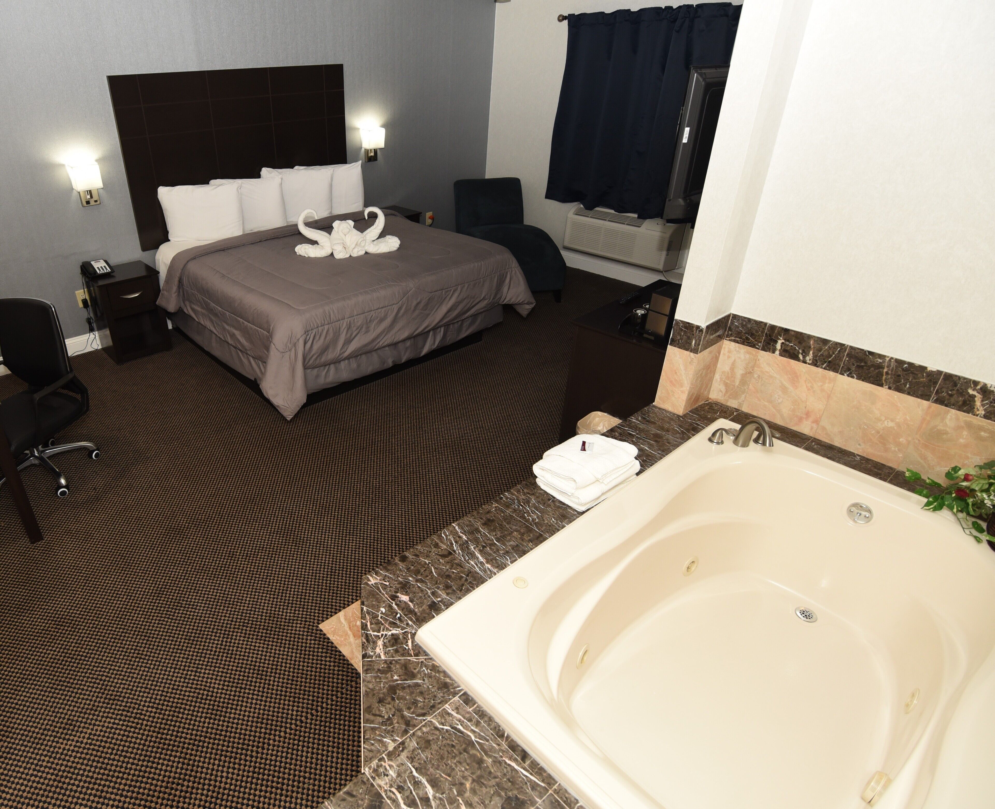 Habitación de hotel con una cama grande, un área de estar independiente, un baño con bañera y una mesita con una silla.