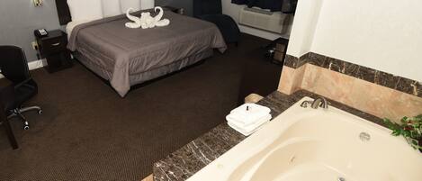 Habitación de hotel con una cama grande, un área de estar independiente, un baño con bañera y una mesita con una silla.