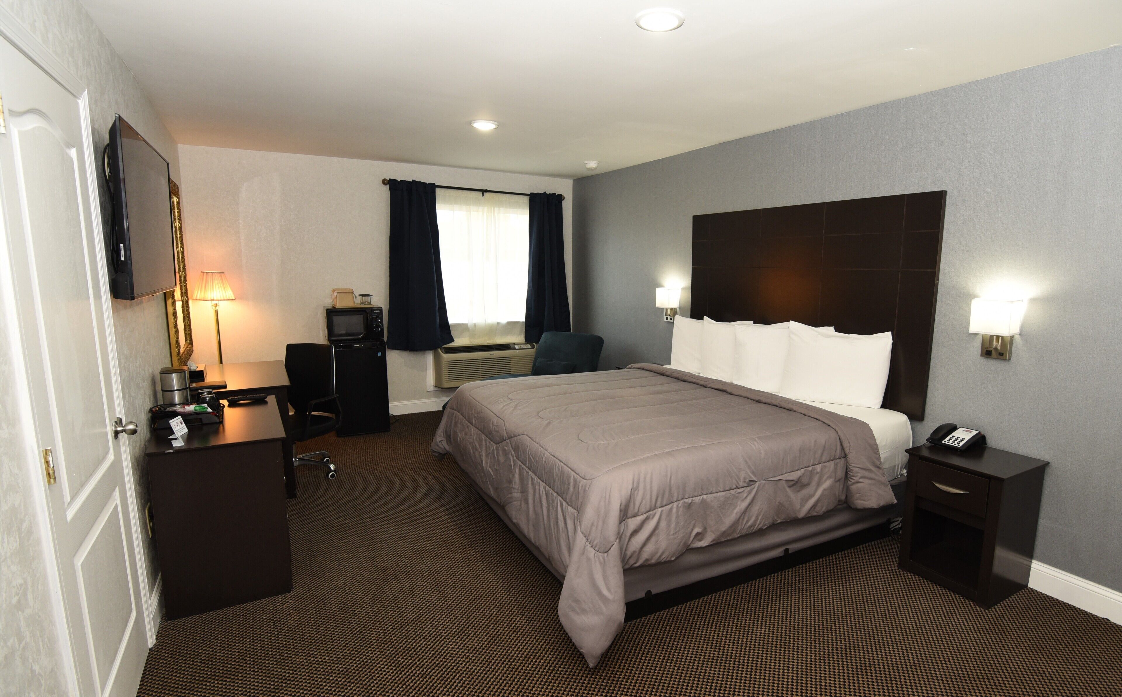 A hotel room with a bed, bedside table, lamp, desk with a computer, and a TV.