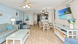 A spacious living room with a ceiling fan, a dining area with a white table and chairs, and a kitchen in the background.