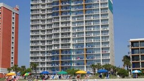 A multi-story hotel with balconies, located near a beach with umbrellas and people.