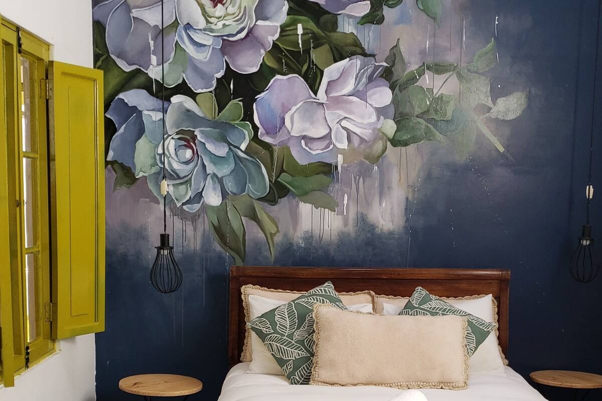 Ein Schlafzimmer mit einem Bett, einer grün-weißen Schachbrettdecke, einem Kopfkissen mit Blätermotiv und einer Wandmalerei mit Blumen.