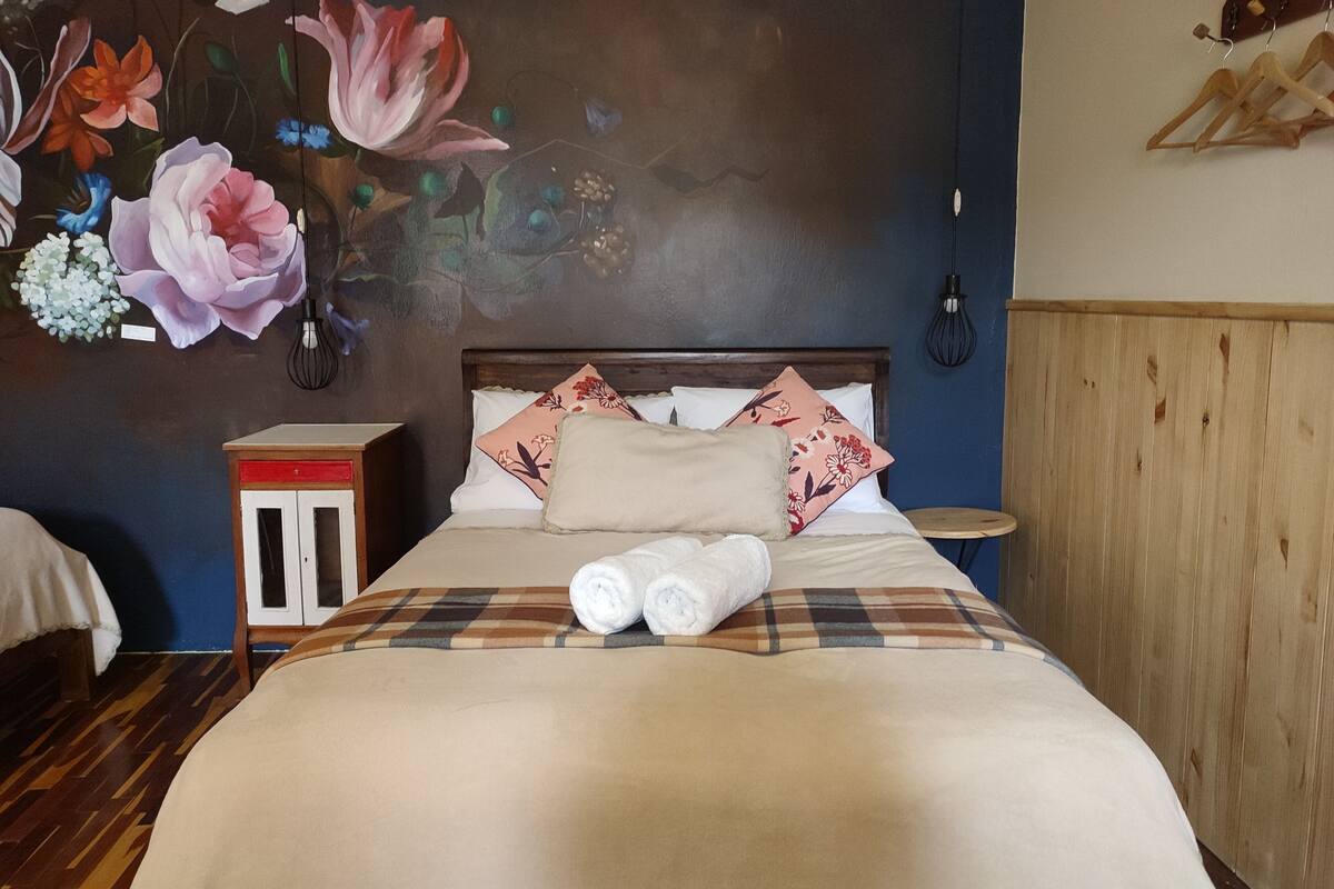 Ein ordentlich bezogenes Bett mit einer karierten Tagesdecke und mehreren Kissen, ein kleiner Nachttisch mit Lampe und eine Wand mit floralem Wandbild.
