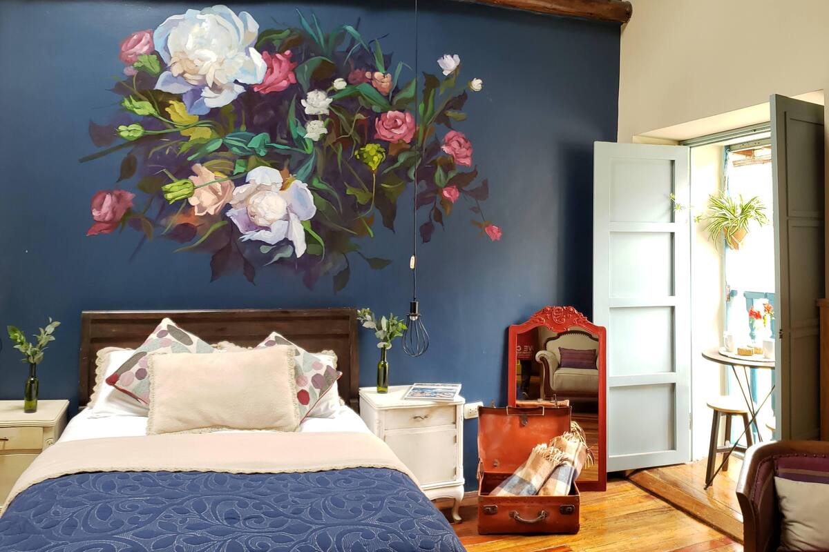 Ein Schlafzimmer mit einer großen Blumenmalerei, einer Holzbalkendecke, einem Bett mit blauer Bettdecke, einem Nachttisch, einem Spiegel und einem Koffer auf dem Boden.