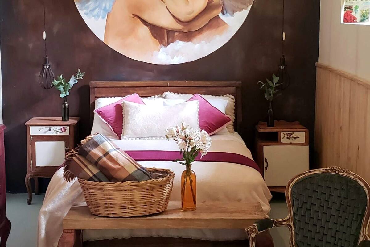 Ein Schlafzimmer mit einem großen, runden Gemälde von Cherubs, einem Bett mit rosa und weißer Bettwäsche, einem Korbwäscheschrank und einem grünen Polsterstuhl.