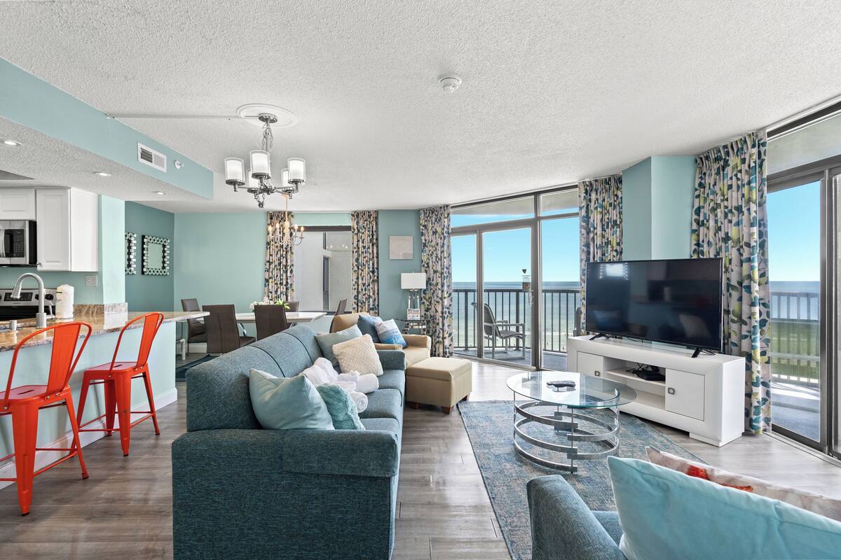 A modern living room with a sofa, ottoman, and TV stand, a dining area with chairs, and a balcony with a view.