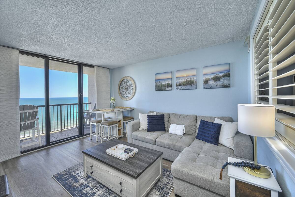 A living room with a large sliding glass door to the ocean, a gray sofa, a coffee table, and a lamp.
