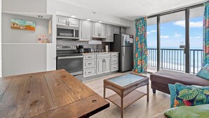 A modern kitchen with white cabinets, a dining table, and a view of the ocean through large windows. - Beach Colony Resort by Elliott Beach Rentals (Myrtle Beach)