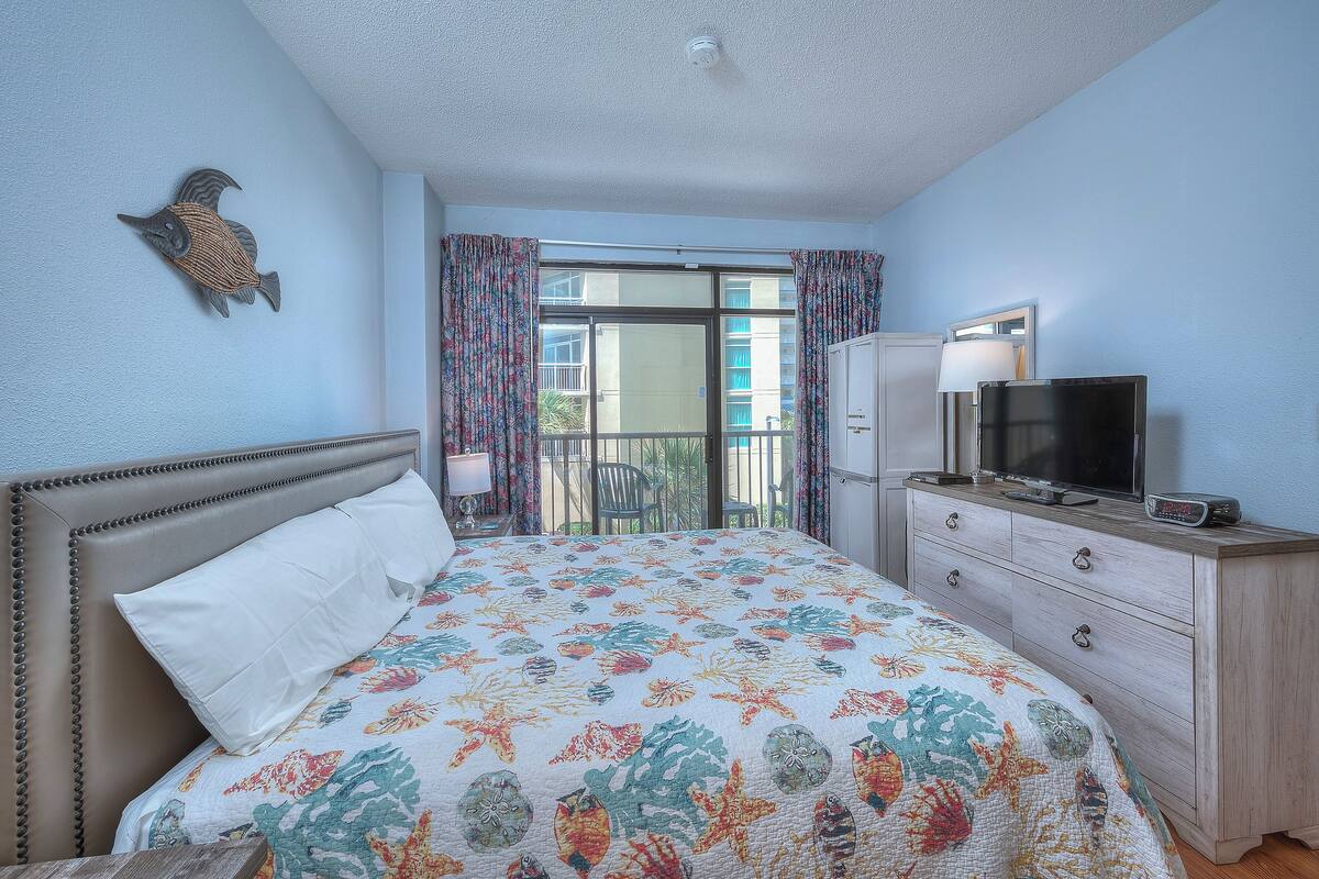 A bedroom with a bed, a dresser, a television, and a balcony with curtains.