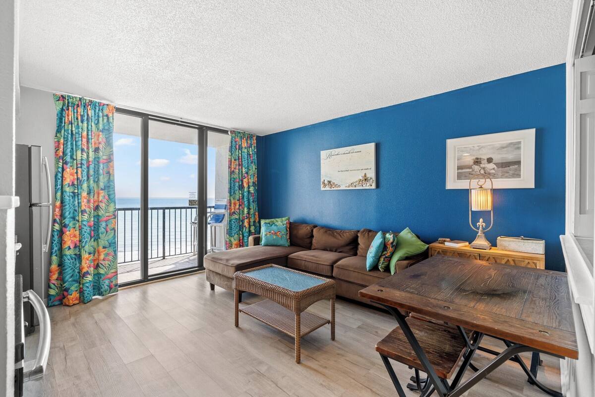 A living room with a sectional sofa, a wooden coffee table, and a view of the sea through large windows.