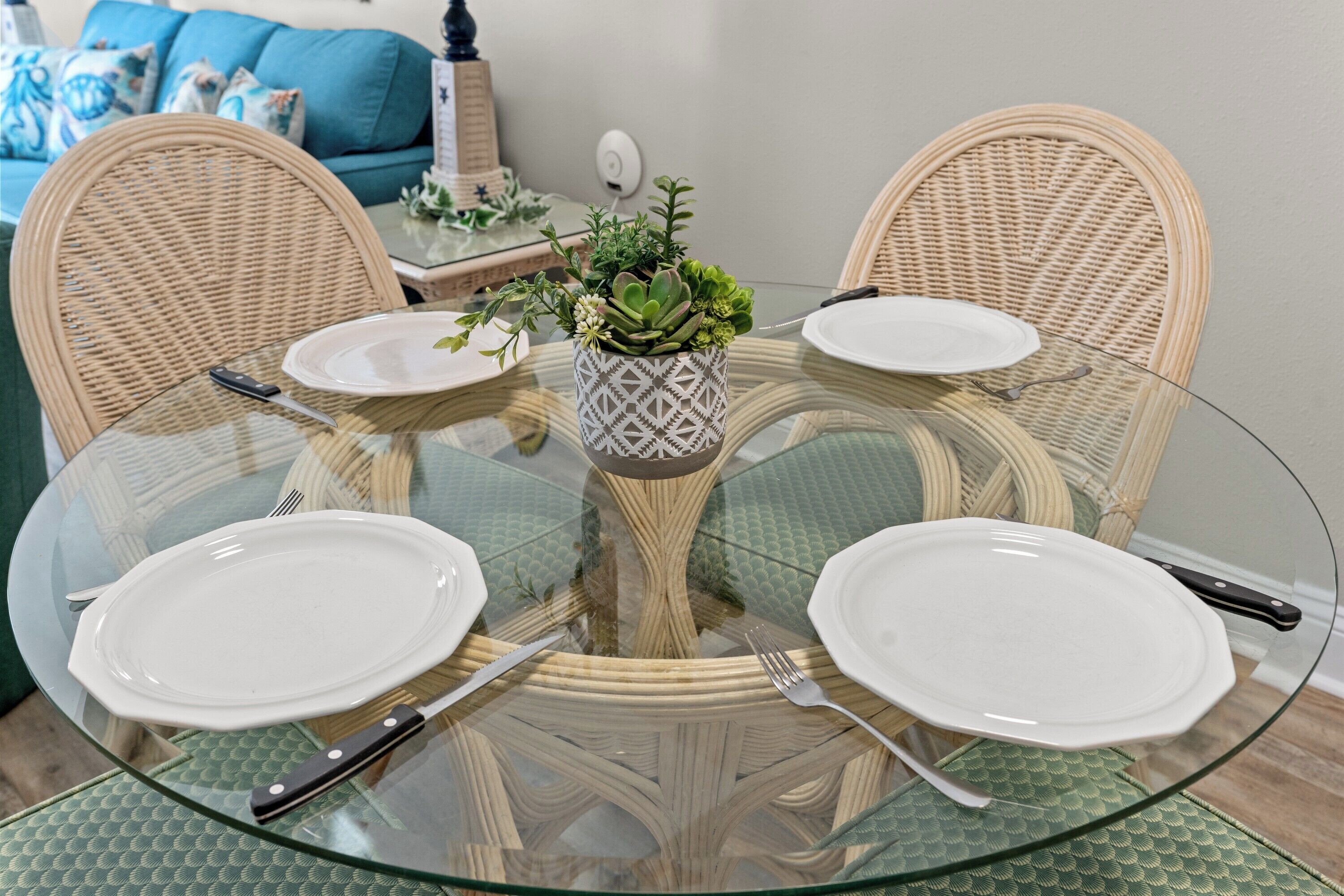 Una mesa de vidrio redonda con cuatro sillas, una planta en maceta en el centro y un plato blanco en cada silla.