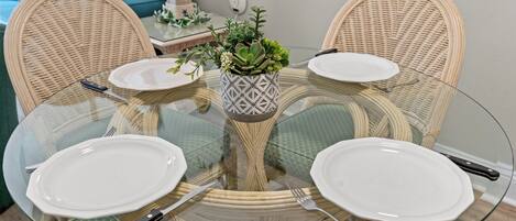 Una mesa de vidrio redonda con cuatro sillas, una planta en maceta en el centro y un plato blanco en cada silla.