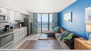 A modern kitchen with white cabinets, a stainless steel refrigerator, and a coffee maker. There is a balcony with a view of the ocean. - Beach Colony Resort by Elliott Beach Rentals (Myrtle Beach)