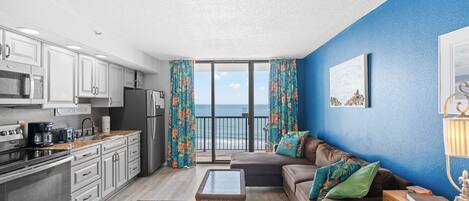 A modern kitchen with white cabinets, a stainless steel refrigerator, and a coffee maker. There is a balcony with a view of the ocean.