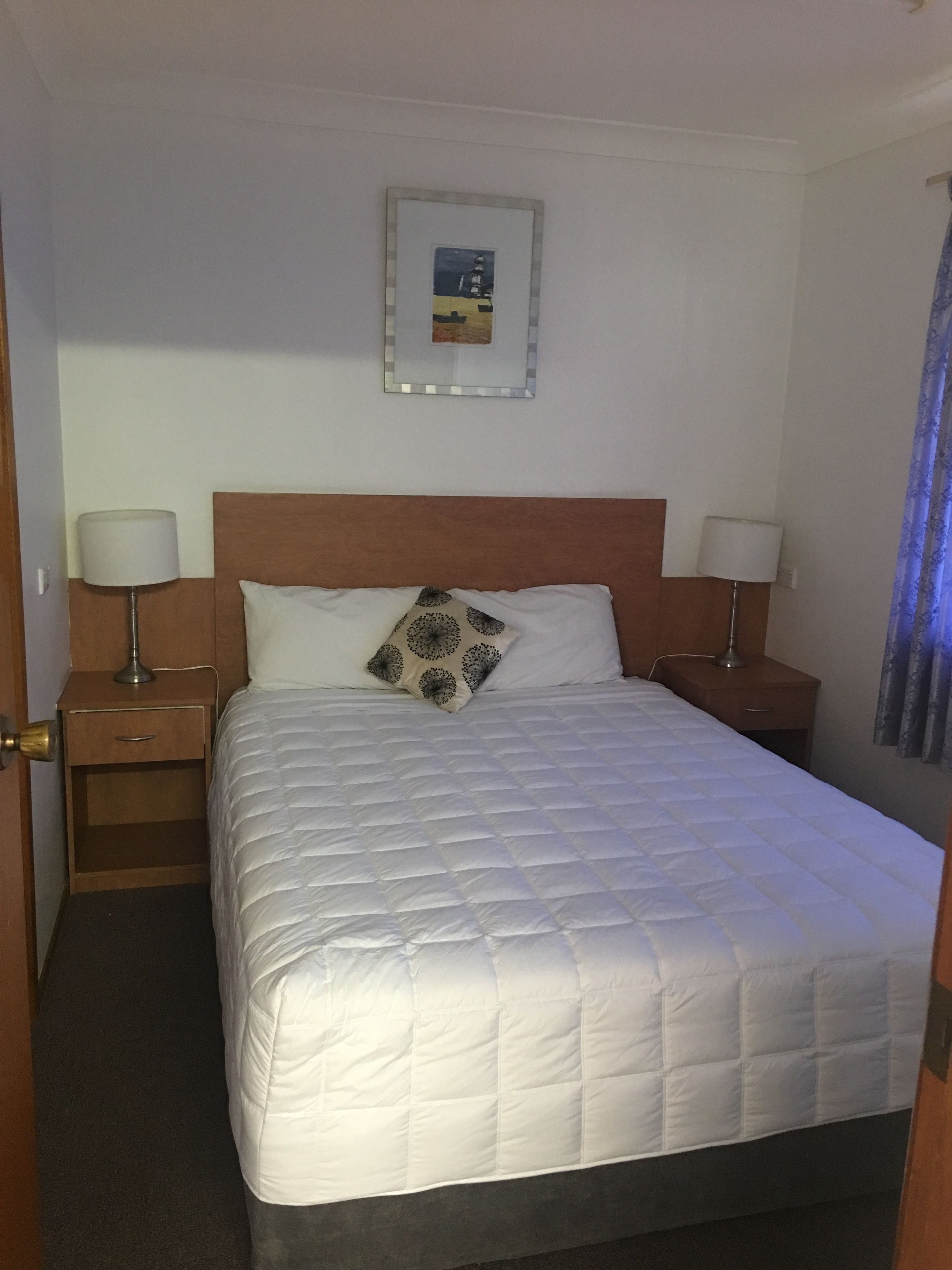 Ein Hotelzimmer mit einem großen Bett, zwei Nachttischen mit Lampen, einem Holzkopfteil, einem gerahmten Bild an der Wand und einer Tür links.