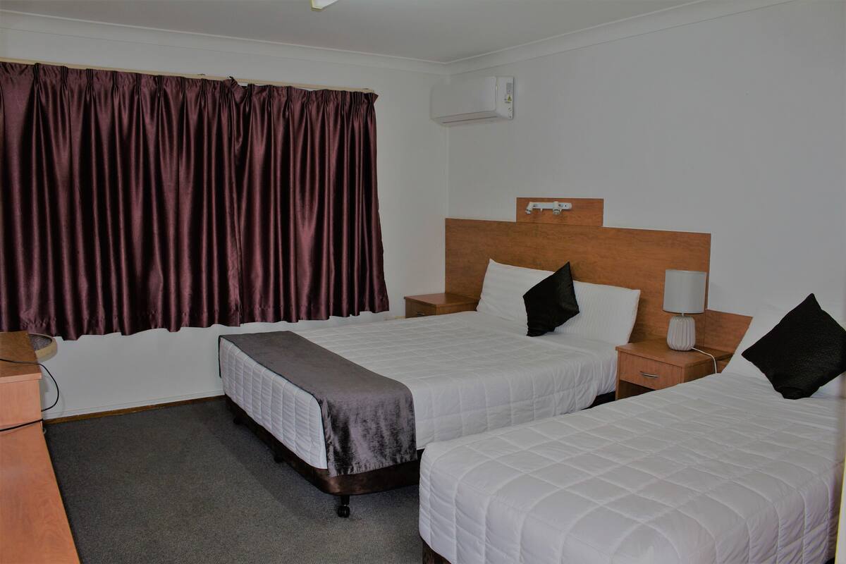 A hotel room with two beds, a wooden headboard, a bedside table with a lamp, and a window with curtains.