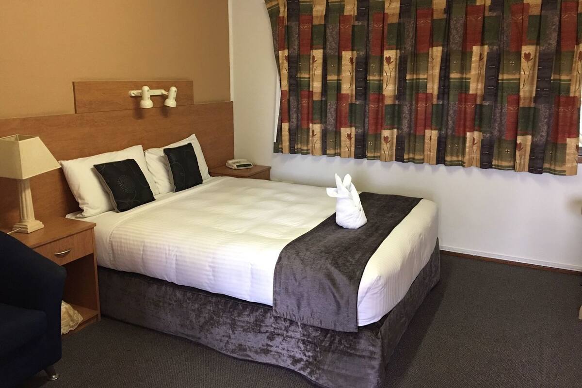A hotel room with a bed, a nightstand, a lamp, a bedside table, a window with patterned curtains, and a wall-mounted clock.
