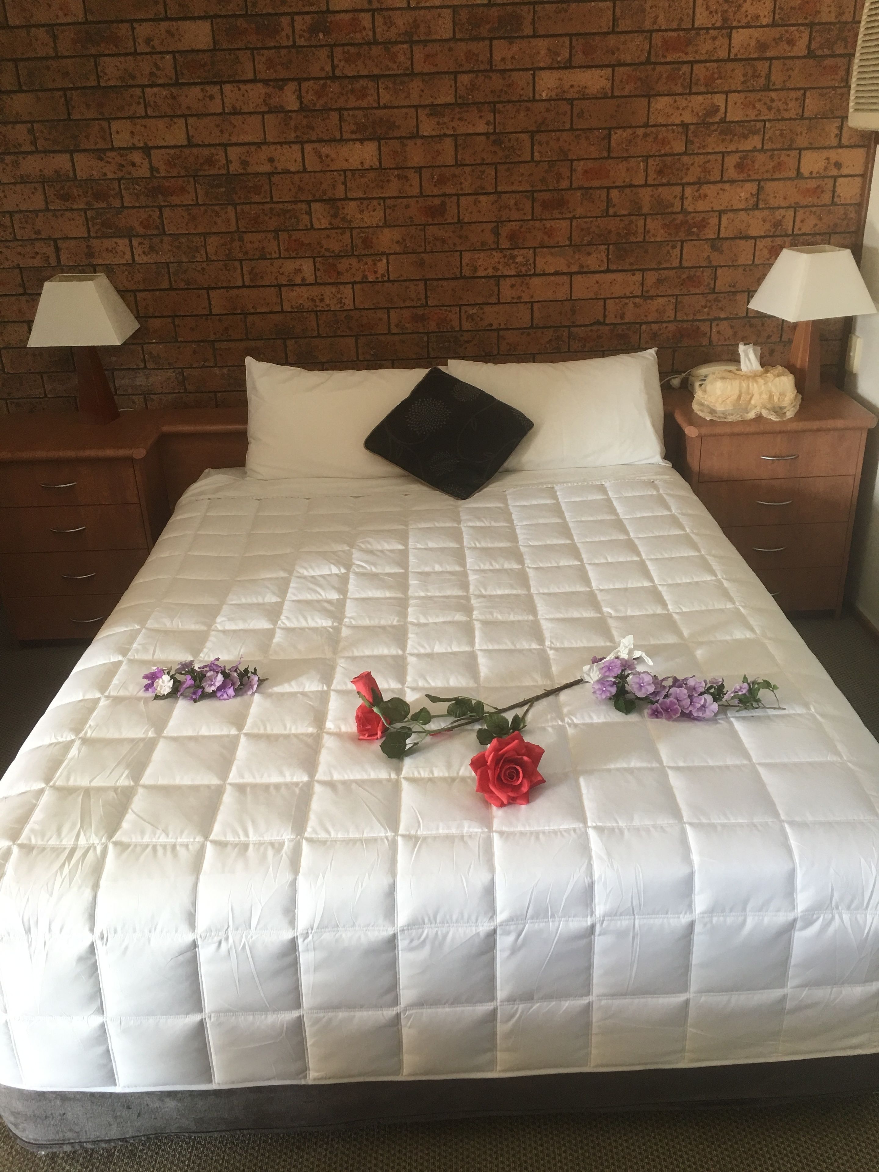 Una camera d'albergo con una parete in mattoni, una coperta bianca trapuntata, un cuscino nero e un bouquet di fiori sul letto.