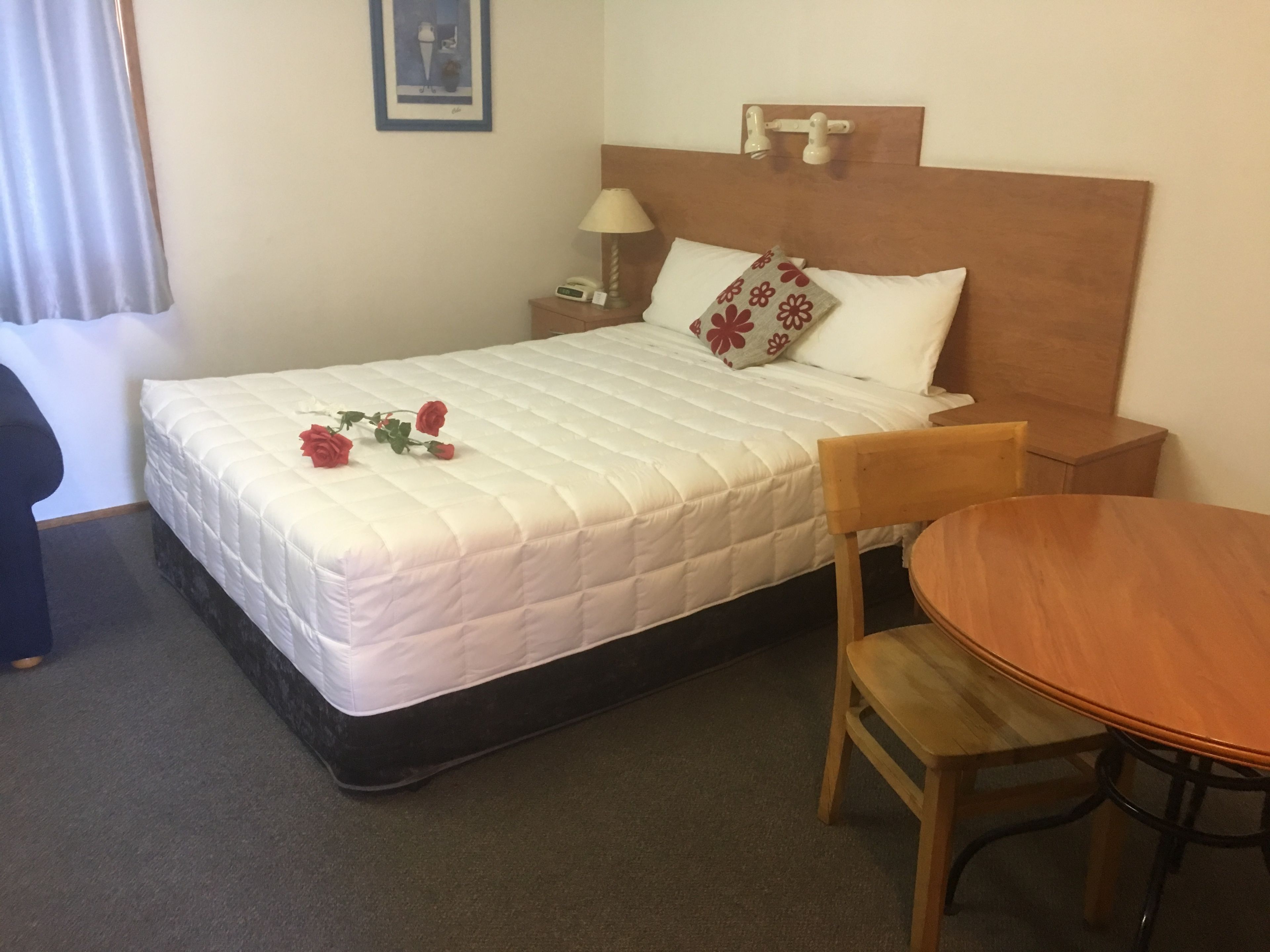 A neatly made bed with a white quilt, a bedside table with a lamp, a wooden chair, and a small round table.