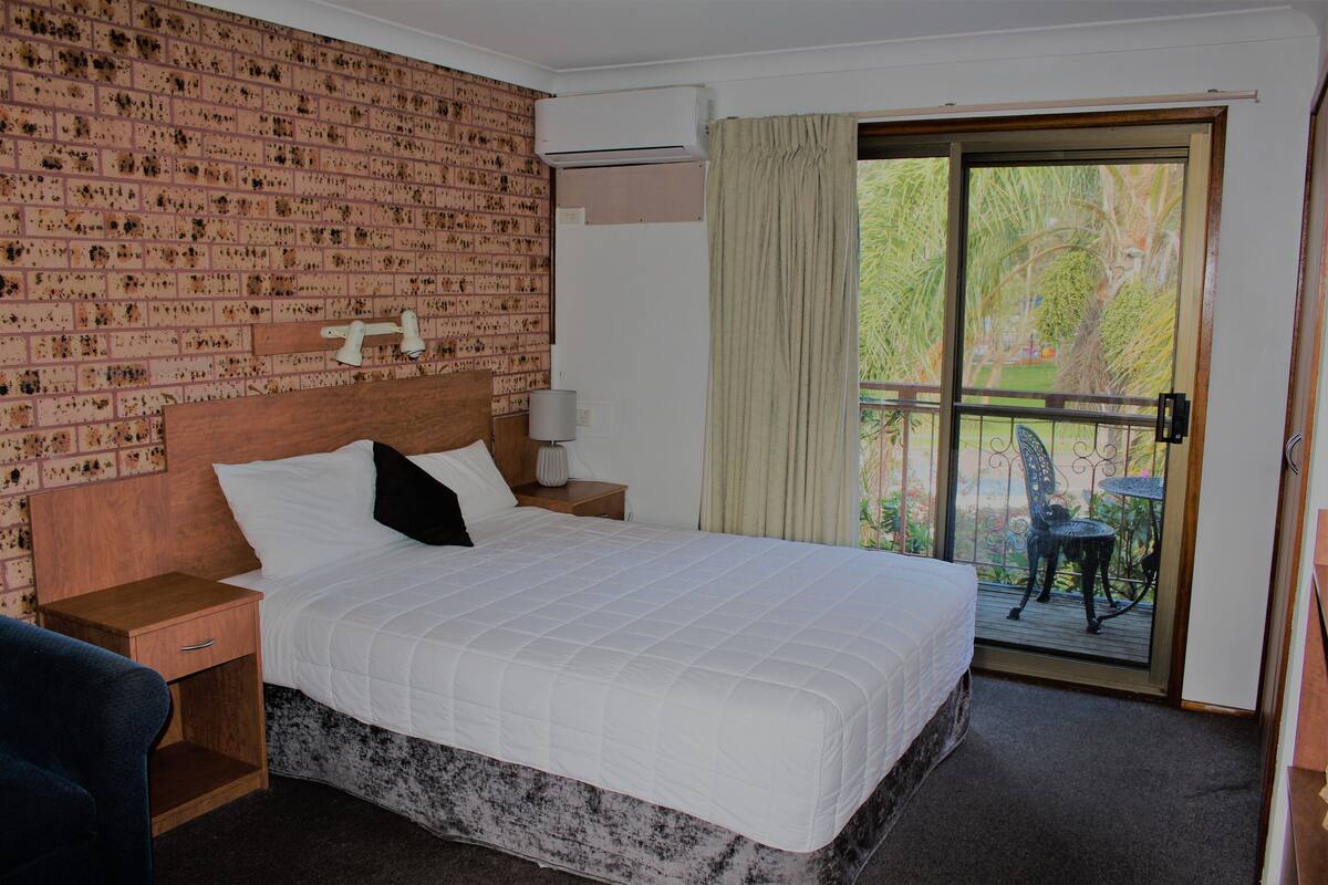 A hotel room with a brick-patterned wall, a large bed with white bedding, a wooden nightstand, a small table with a lamp, and a view of a balcony with outdoor furniture.