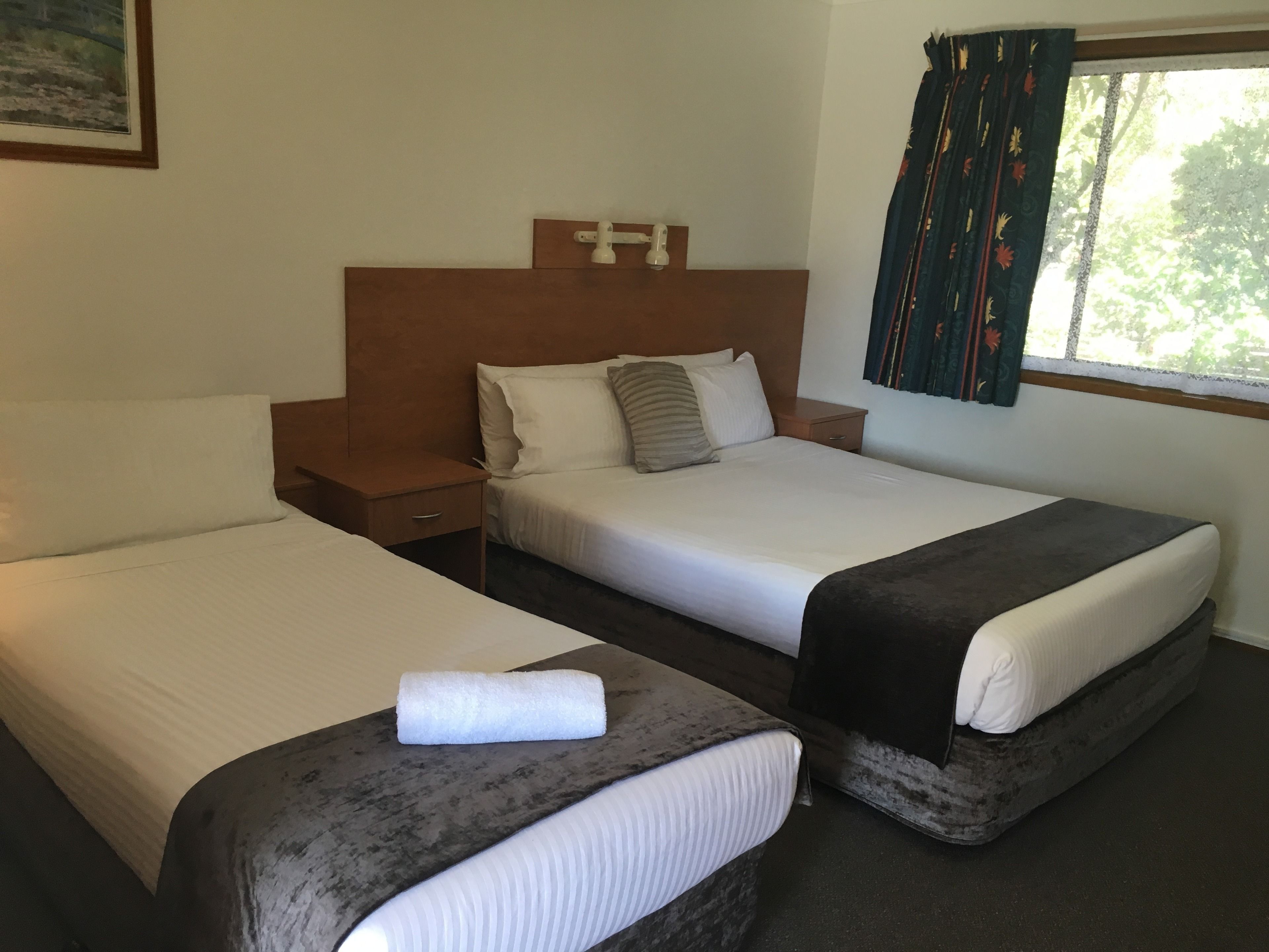 A hotel room with two single beds, a wooden headboard, a small bedside table, and a window with curtains.