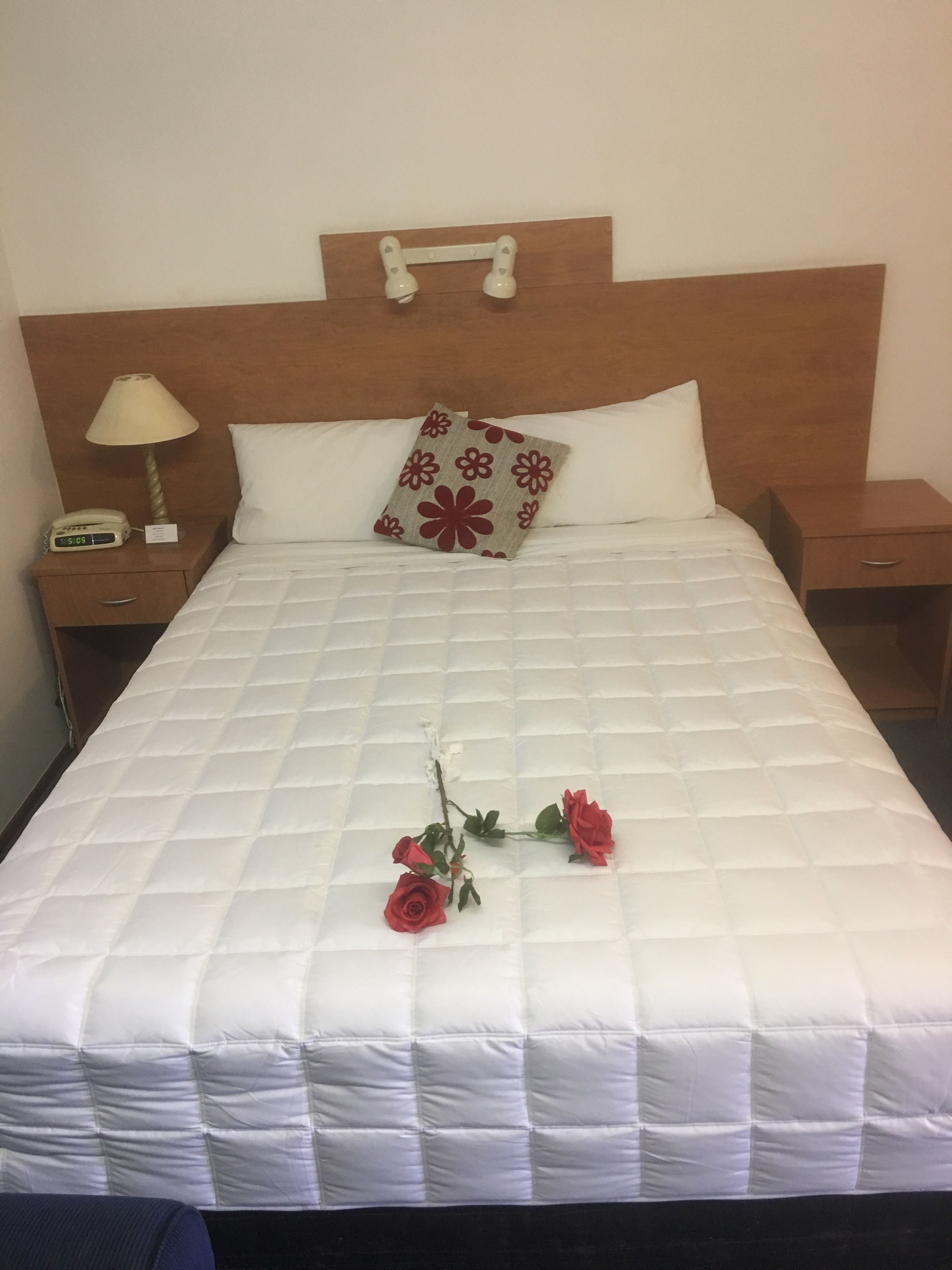 A neatly made bed with a white quilt, a wooden headboard, a bedside lamp, and a nightstand with a clock.
