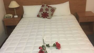A neatly made bed with a white quilt, a wooden headboard, a bedside lamp, and a nightstand with a clock.