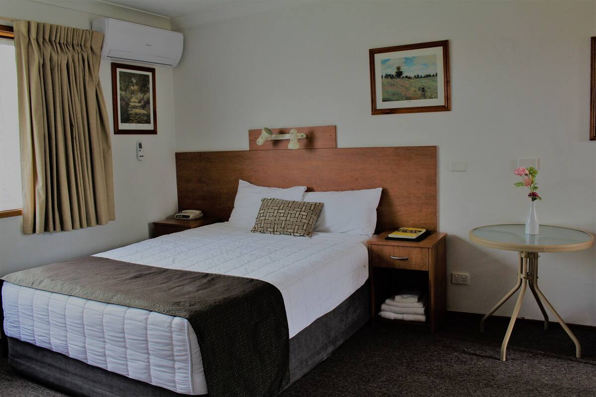 A hotel room with a bed, bedside tables, a small round table, a wall-mounted air conditioner, and a painting.