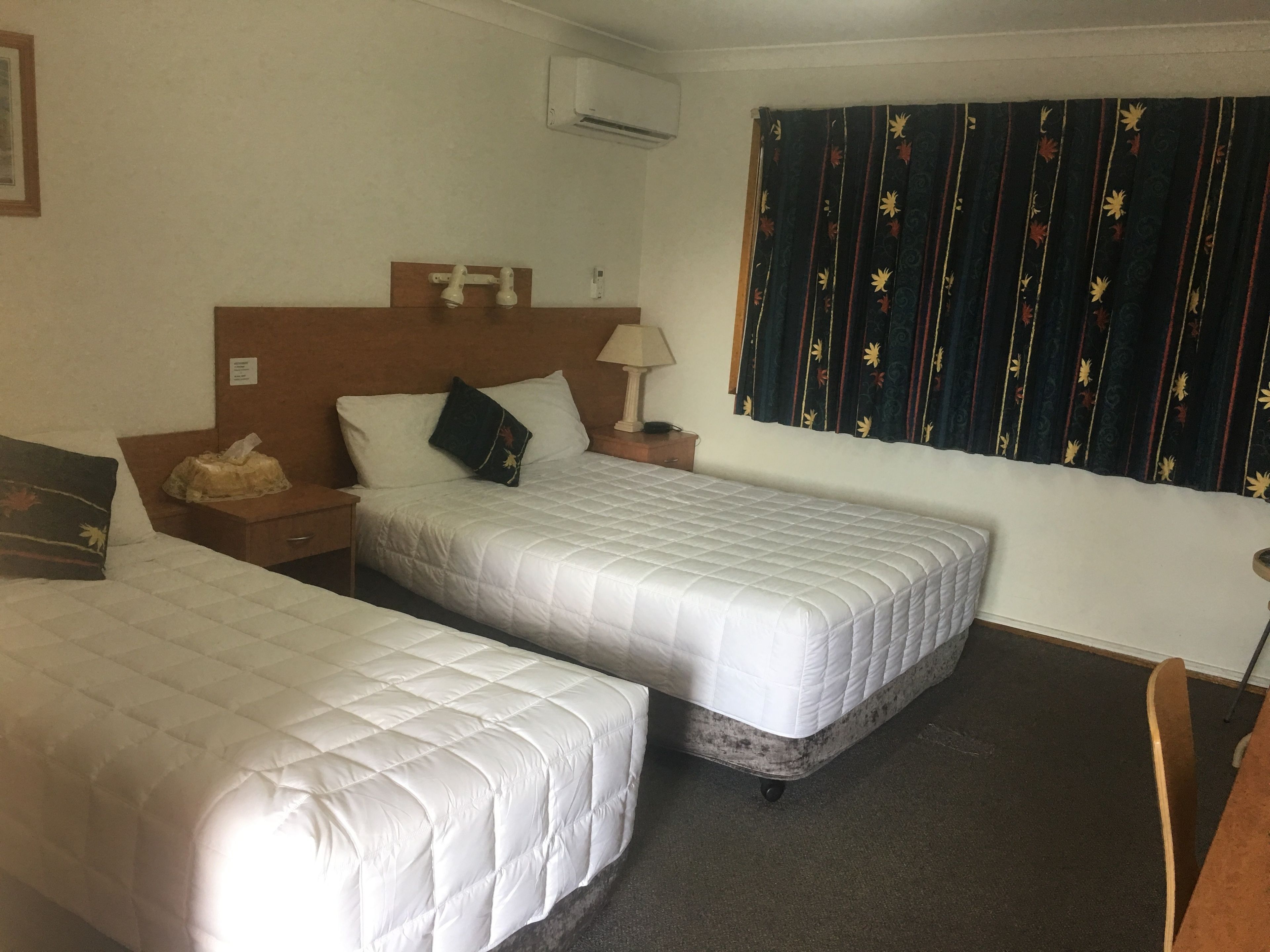A hotel room with two single beds, a wooden headboard, a nightstand with a lamp, a small table, and a window with patterned curtains.