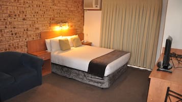 A hotel room with a brick-patterned wall, a bed with a white and brown bedspread, a blue armchair, a wooden nightstand, a desk with a computer, and a window with curtains.