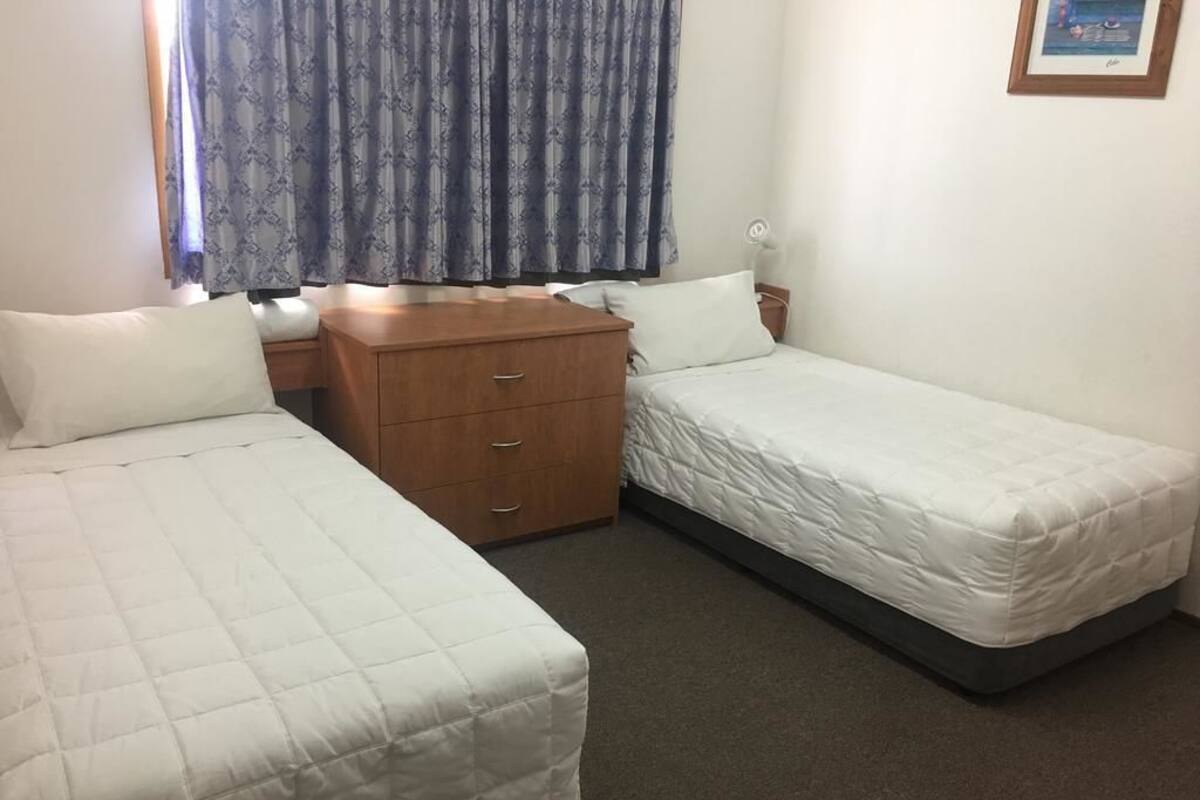 A small, clean room with two single beds, a wooden dresser, and a window with patterned curtains.