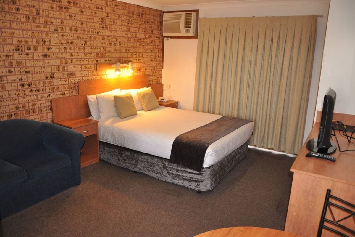 A hotel room with a brick-patterned wall, a bed with white bedding and a brown throw, a desk with a computer, and a blue armchair.