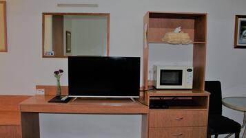 A hotel room with a wooden desk, a flat-screen TV, a microwave, a small shelf with a decorative item, a chair, and a vase with flowers.
